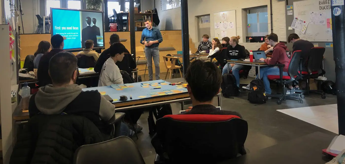 Marco Kramer geeft les aan een groep studenten van NHL Stenden; op het scherm staat ‘First: you need focus’ en op tafel liggen post-its.